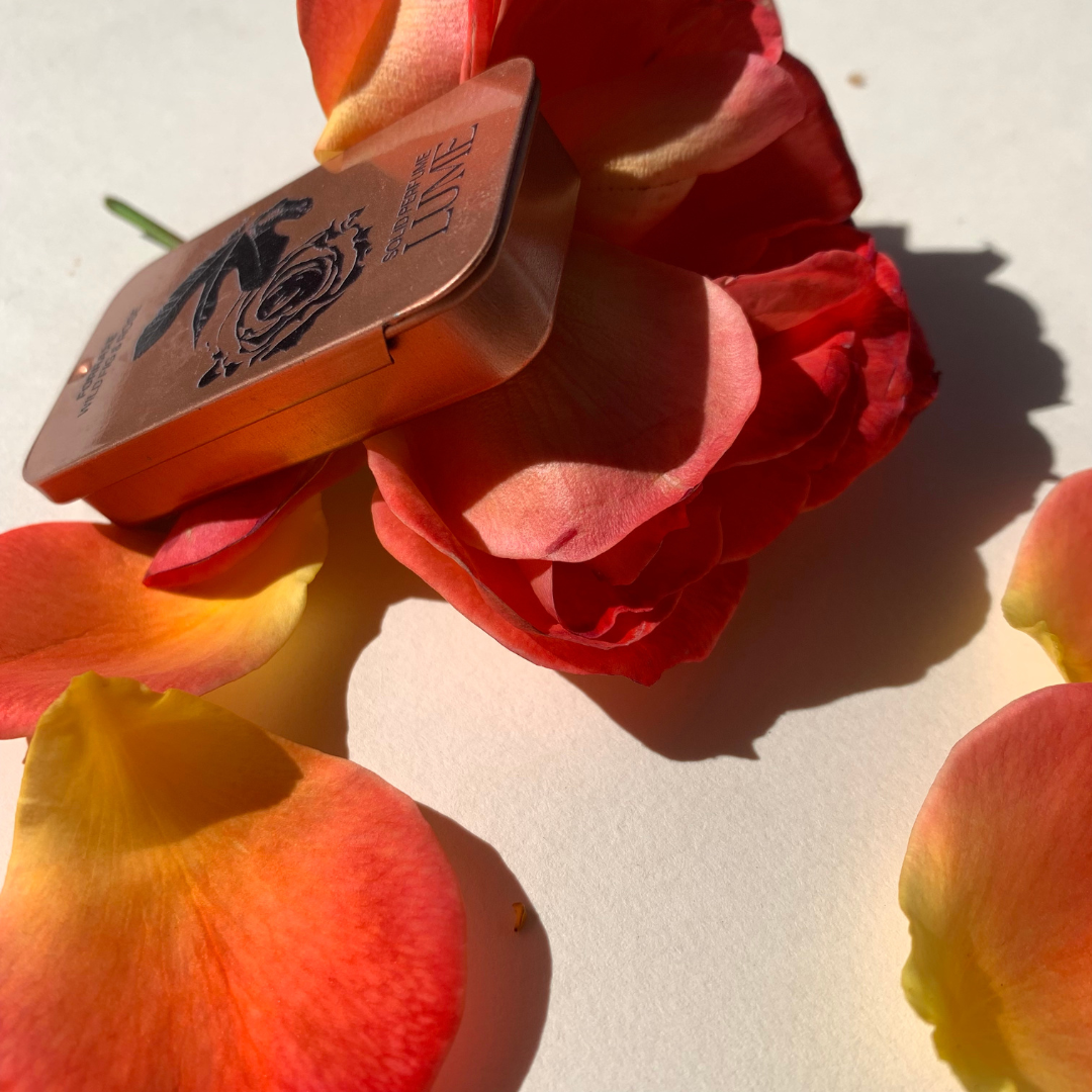 Side view of a solid perfume in a metal tin, resting on rose petals.