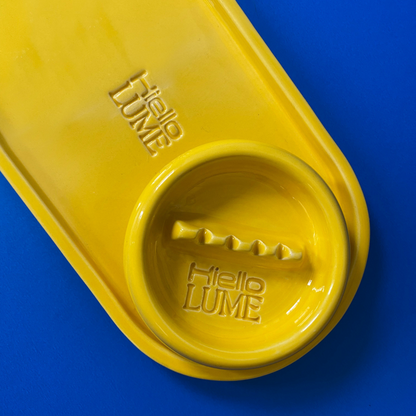 A closeup of a ceramic tray and matching ashtray, embossed with the logos of Hiello and LUME.